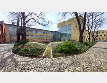 Innenhof eines modernen Gebäudekomplexes mit gelben Fassaden und großen Glasflächen. Der Hof ist von Kopfsteinpflasterwegen, Bäumen und gepflegten Grünflächen mit niedrigen Pflanzen gesäumt.