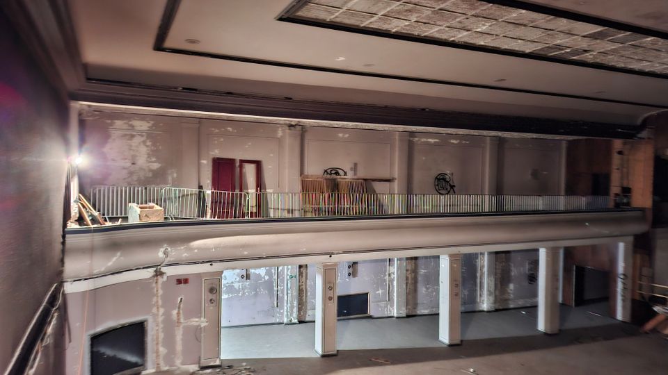 Blick in den Innenraum eines verlassenen, zweigeschossigen Gebäudes mit umlaufender Empore und niedriger Brüstung. Abgeblätterte Wandflächen, Säulen, offene Türrahmen und teilweise beschädigte Deckenplatten prägen den Raum.