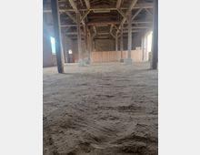 Ein leerer, großer Innenraum mit sandigem Boden, Holzbalken als tragende Struktur und einer offenen Dachkonstruktion. Die Wände bestehen aus unverputzten Ziegeln, und im Hintergrund sind Holzwände sowie kleine Fenster sichtbar.