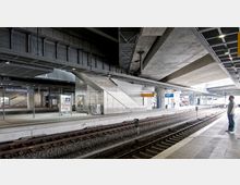 Moderne Bahnhofshalle des Berliner Südkreuzes mit mehreren Gleisen, Überdachungen und Betonarchitektur. Im Bild sind Beschilderungen, Treppen, Informationsanzeigen und ein wartender Passant auf dem Bahnsteig zu sehen.