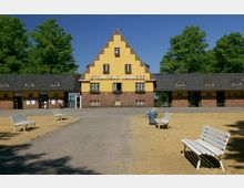 Historisches Eingangsgebäude des Strandbads Wannsee mit gelb gestrichenem, giebelständigem Hauptbau und rotbrauner Ziegelfassade, flankiert von niedrigeren Gebäudeflügeln mit kleinen Rundbogenfenstern. Im Vordergrund ein sandiger Platz mit weißen Holzbänken und einer grünen Baumkulisse im Hintergrund.