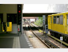 Eine U-Bahn-Haltestelle mit einem gelben Zug auf der rechten Seite, mehreren Gleisen, Signalanlagen und einem Spiegel an einem Gebäudeteil auf der linken Seite. Im Hintergrund führt die Überdachung der Station zu weiteren Gleisverzweigungen und Lichtsignalen.