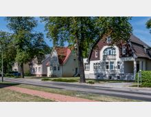 Straßenszene mit einer Reihe von Einfamilienhäusern im traditionellen Stil, teils mit roten Schrägdächern und verputzten Fassaden. Im Vordergrund befindet sich eine Straße mit Gehwegen und Bäumen, die Schatten spenden.