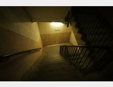 Treppenhaus in einem Gebäude mit herunterführenden Betonstufen, metallischem Geländer und gelblicher Beleuchtung. An der Wand ist das Wort „Untergeschoss“ zu erkennen.