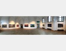 Panoramablick in einen Ausstellungsraum mit hohen weißen Wänden und Spitzbogenfenstern. Die Wände zeigen großformatige Gemälde, während der Raum durch gotische Gewölbedecken und einen hölzernen Boden geprägt ist.