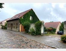 Dorfstraße mit Kopfsteinpflaster, gesäumt von Häusern mit roten Ziegeldächern; ein Haus ist vollständig mit Efeu bewachsen, daneben ein kleiner Vorgarten mit Blumen. Im Hintergrund sind weitere Häuser und ein geparktes Auto sichtbar.