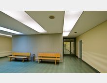 Ein schlichter Flur in einem Gebäudeeingangsbereich mit grünem Boden und beigefarbenen Wänden. Zwei Holzsitze und ein Mülleimer stehen an der linken Wand, während sich am Ende des Gangs eine Glastür mit der Aufschrift "Ausgang" befindet.