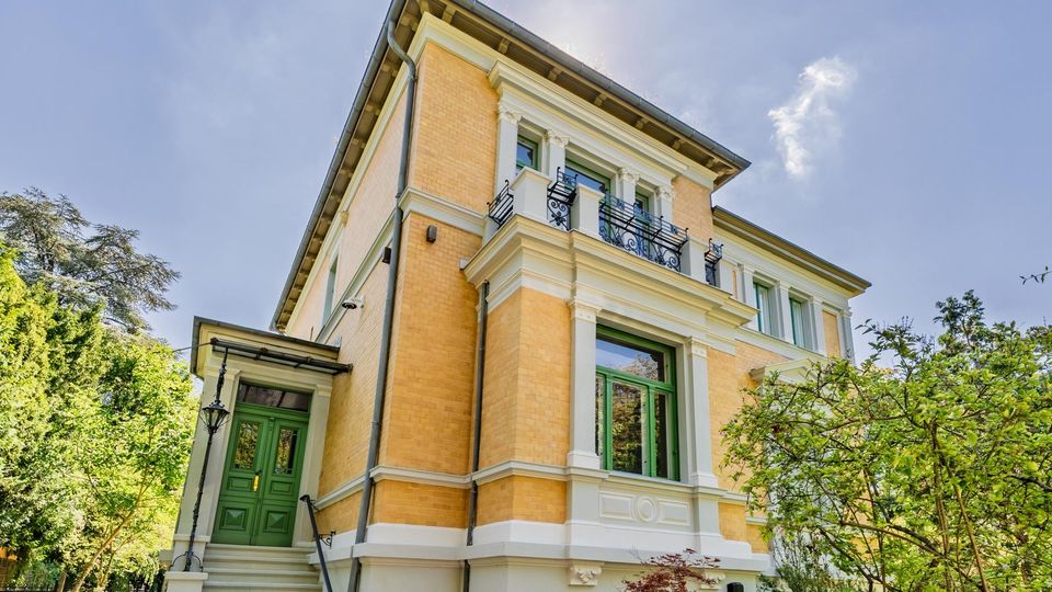 Ansicht einer mehrstöckigen Gründerzeit-Villa aus gelbem Backstein mit weißen Stuckelementen, grün gerahmten Fenstern und kleinem Balkon mit schmiedeeisernem Geländer. Links führt eine Treppe zu einer grünen Eingangstür, umgeben von Gartenbäumen und Sträuchern.