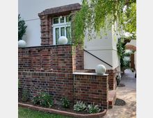 Fassade eines Wohnhauses mit weiß verputzten Wänden und roten Klinkersteinen. Im Vordergrund eine kleine Terrasse mit gemauerten Brüstungen, dekoriert mit weißen Kugelleuchten, umgeben von einem Blumenbeet und einem gepflasterten Weg.