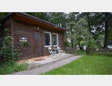 Kleines Holzhaus mit einfacher Bauweise, bestehend aus einer Holztür und zwei weißen Fenstern. Davor stehen ein kleiner Tisch und zwei Klappstühle auf einer gepflasterten Terrasse, umgeben von grüner Wiese und Bäumen.