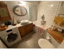 Kleines Badezimmer mit weiß gefliesten Wänden, gemusterten Bodenfliesen und Holzdetails. Eine freistehende Waschmaschine, ein Waschtisch mit ovalem Spiegel, eine Badewanne und ein Handtuchheizkörper sind sichtbar.