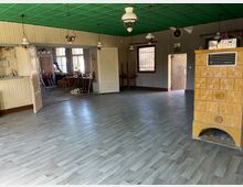 Ein leerer Raum mit grauem Laminatboden und einer grünen Decke, ausgestattet mit mehreren hängenden Lampen. In der rechten Ecke befindet sich ein gelber Kachelofen, und im Hintergrund sind zwei geöffnete Türen, die zu einem angrenzenden Raum mit Fenstern und Möbeln führen.