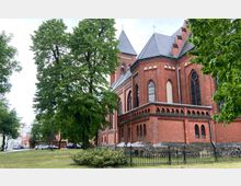 Backsteinkirche im neugotischen Stil mit spitzen Türmen und großen, spitzbogigen Fenstern, umgeben von Bäumen und einer Grünfläche; im Vordergrund ein schmiedeeiserner Zaun.