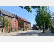 Straßenszene mit historischen Backsteingebäuden auf der linken Seite, darunter zwei zweigeschossige Häuser mit Satteldächern und weißen Fensterrahmen. Rechts säumen Bäume den Gehweg, dahinter vereinzelt moderne Häuser und ein kleines Gebäude mit dunklem Dach.