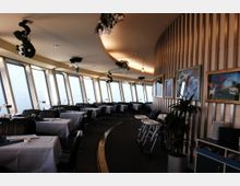 Ein modern eingerichtetes Restaurant mit großen, geneigten Fensterfronten, die eine Aussicht auf die Umgebung ermöglichen. Die Tische sind mit weißen Tischdecken gedeckt, dekoriert mit Pflanzen und Gemälden an einer Holzvertäfelung an der Wand.