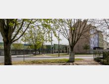 Straßenszene mit Bäumen im Vordergrund, einem Backsteingebäude rechts und einer Mauer mit darüber verlaufenden Bahngleisen im Hintergrund. Ein Straßenschild weist auf die "Franz-Schubert-Straße" hin, daneben ist ein geparktes Auto sichtbar.