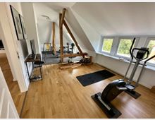 Dachgeschossraum mit Holzboden und Fitnessgeräten, darunter ein Crosstrainer, eine Hantelbank und ein Rudergerät; sichtbare Dachschrägen, freiliegende Holzbalken und große Fenster, die viel Tageslicht hereinlassen.