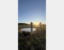 Auf einer offenen Wiese bei Sonnenuntergang stehen zwei Personen neben einer Wasserpfütze, während ein Hund mit Geschirr im Vordergrund des Bildes zu sehen ist. Im Hintergrund erstrecken sich Bäume am Horizont.