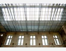 Raum mit Ziegelwänden und rechteckigen Fenstern, kombiniert mit einer modernen Glas-Stahl-Decke, die durch ein Gittermuster strukturiert ist und viel Tageslicht einlässt.