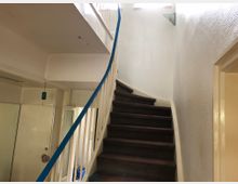 Treppenhaus in einem Wohngebäude mit dunkelbrauner, gewendeter Treppe und weiß gestrichenen Wänden. Ein blaues Handlaufband verläuft entlang des Treppengeländers, und im unteren Bereich befinden sich weiße Türen.