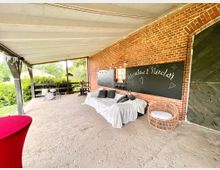 Überdachter Außenbereich mit Ziegelfassade, einem schwarzen Holzschild mit den Namen „Marielena & Nikolai“, einem Sofa mit weißen Bezügen, einem Korbstuhl und einer kleinen Trommelbühne. Im Hintergrund sind grüne Hecken und eine Wiese zu sehen.