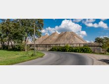 Eine kurvige Asphaltstraße führt an einer Anlage mit hohen Kieshaufen vorbei, die hinter einer Betonmauer liegen. Im Vordergrund befinden sich Grasflächen und Bäume, während der Himmel mit einigen Wolken bedeckt ist.
