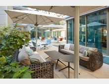 Moderner Innenhof mit einer Terrasse, ausgestattet mit geflochtenen Sitzgruppen mit Kissen, kleinen Tischen, Sonnenschirmen und bunten Stühlen. Im Hintergrund sind Glasfassaden und eine Begrünung sichtbar.