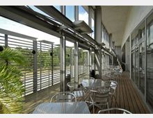 Ein moderner Balkon mit Holzfußboden, ausgestattet mit silbernen Metalltischen und -stühlen. Große Glasfenster und eine Lamellenwand bieten Blick auf grüne Bäume im Außenbereich.