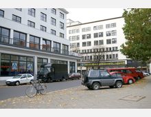 Straßenszene vor einem modernen weißen Gebäude mit mehreren Etagen und großen Fenstern. Im Vordergrund sind parkende Autos, ein schwarzer Lieferwagen, ein Fahrrad sowie Laub auf der Straße; auf einem Schild ist "Neues Berliner Kabarett-Theater" zu lesen.