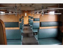 Innenraum eines historischen Zugabteils mit hölzernen Trennwänden und grünen Sitzpolstern, ausgestattet mit Gepäckablagen aus Metall über den Sitzen. Am Ende des Gangs befindet sich eine Holztür mit einem kleinen Fenster.