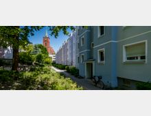 Wohnsiedlung mit pastellfarbenen Mehrfamilienhäusern, einem Gehweg und Büschen im Vordergrund. Im Hintergrund ist eine rote Backsteinkirche mit Turm und grünem Spitzdach zu sehen, umgeben von Bäumen.