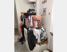 A bathroom with a white bathtub being used as a drying area for clothes, featuring a portable drying rack filled with garments. Above the tub is a wall-mounted water heater, and recessed shelves contain personal care products.