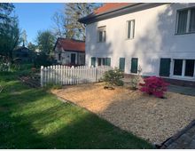 Großer Garten mit einem weißen, zweistöckigen Wohnhaus mit grünen Fensterläden im Hintergrund. Im Vordergrund ein mit Holzschnitzeln bedecktes Beet, das von einem weißen Holzzaun umgeben ist, und eine pink blühende Pflanze. Rechts und links stehen Bäume und Büsche im grünen Rasenbereich.