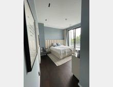 Helles Schlafzimmer mit einem großen Bett, gepolstertem Kopfteil und weißer Bettwäsche, flankiert von zwei Nachttischen. Der Raum hat eine Glas-Balkontür mit Vorhängen, die Tageslicht hereinlässt, dunklen Holzboden und eine schlichte, moderne Einrichtung.