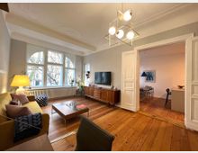 Wohnzimmer in einem Altbau mit großen Bogenfenstern, Holzboden und Stuckdecke. Der Raum ist mit einer gelben Couch, Holzmöbeln, einem Fernseher und einer modernen Hängelampe eingerichtet; im Hintergrund führt eine Doppeltür in einen Essbereich.