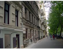 Straßenansicht mit Altbauten auf der linken Seite, die Fassaden zeigen dekorative Stuckdetails und teils sichtbare Abnutzung. Rechts säumen Bäume den Gehweg, wo einzelne Passanten spazieren, und parkende Autos sind im Schatten der Bäume sichtbar.