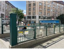 Eingang zur U-Bahn-Station „Weinmeisterstraße“ in einer städtischen Umgebung, umgeben von mehrstöckigen Wohn- und Geschäftsgebäuden. Der Eingang ist durch ein blaues Schild mit Stationsnamen gekennzeichnet und von grünen Metallgeländern umgeben.