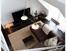 Wohnzimmer in einer Dachgeschosswohnung mit dunklem Holzboden, einer großen braunen Ledercouch, weißen Vorhängen vor einem großen Fenster, einem flachen Fernseher an der Wand und einer Hängelampe mit zwei Leuchten. Im Raum befinden sich ein Korbsessel, ein Kaktus in einem Terrakottatopf und ein heller Teppich.