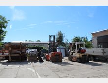Industriegelände im Freien mit zwei Gabelstaplern im Vordergrund, mehreren Metallgeräten und Baumaterialien sowie einem grauen Gebäude und Bäumen im Hintergrund unter blauem Himmel.