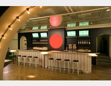 Eine moderne Bar mit gemustertem Tresen und schwarzen Barhockern auf einem Holzboden. Die Wand im Hintergrund ist dunkel mit einem großen roten Kreis als Dekoration, umgeben von Regalen mit Flaschen und Gläsern sowie mehreren Monitoren.