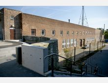 Backstein-Gebäude mit rechteckigen Fenstern in einem Industrie- oder Gewerbegebiet. Im Vordergrund sind eine Treppe mit Metallgeländer, eine Betonmauer und gepflasterte Wege zu sehen, im Hintergrund stehen ein Sendemast und verschiedene technische Anlagen.