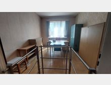 Ein einfacher, spärlich eingerichteter Raum mit Holzfußboden; im Hintergrund steht ein Tisch mit drei Stühlen vor einem Fenster mit halbtransparentem Vorhang. Rechts befinden sich ein hoher Schrank und eine Holzkommode, links ein kleiner Tisch und Regale; im Vordergrund sind Metallgeländer zu sehen.