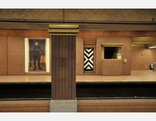 U-Bahn-Station mit Backsteinwänden, einem markanten gemusterten Türdesign in Schwarz-Weiß und einem großformatigen Porträtgemälde an der Wand. Im Vordergrund sind ein Gleis und eine Säule mit brauner Verkleidung zu sehen.
