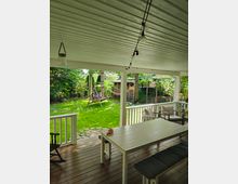 Überdachte Holzveranda mit weißen Geländern und einer Sitzgruppe aus Tisch, Bank und Stühlen, dahinter ein Garten mit Rasen, Bäumen, einer Schaukel, einer Hängematte und einem Holzlager. Lichterketten hängen unter der Decke der Veranda.