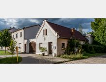 Ein kleines, weißes Einfamilienhaus mit rotem Satteldach und einem Kamin, flankiert von Pflanzen und Sträuchern. Daneben verläuft ein gepflasterter Gehweg, und im Hintergrund sind weitere Gebäude und Bäume erkennbar, unter einem dramatischen, wolkigen Himmel.