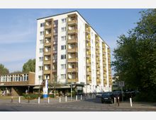 Mehrstöckiges Wohngebäude mit Balkonen, teils gelb abgesetzt, an einer Straßenkreuzung. Im Erdgeschoss befinden sich Geschäfte, darunter ein Blumenladen, und daneben ein niedriges, flaches Gebäude mit Schaufenstern. Im Vordergrund stehen parkende Autos, und rechts ist dichter Baumbestand sichtbar.