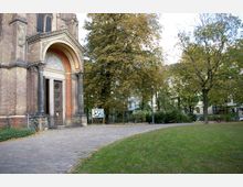 Backsteinfassade einer Kirche mit einem großen, bogenförmigen Eingangsportal links im Bild, daneben eine gepflasterte Fläche, die in eine grüne Parkanlage mit Bäumen und Rasen übergeht. Im Hintergrund sind Wohnhäuser und eine Infotafel sichtbar.