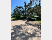 Ein Parkplatz mit sandigem Untergrund, umgeben von hohen Bäumen und Schattenwurf durch das Sonnenlicht. Im Hintergrund steht ein schwarzes Auto, und rechts ist ein teilweise sichtbares Holzgebäude.