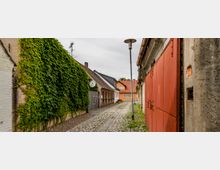 Schmale Gasse mit Kopfsteinpflaster, flankiert von Gebäuden mit unterschiedlichen Fassaden, darunter eine mit Efeu bewachsene Wand und eine große rote Metalltür. Im Hintergrund stehen niedrige Häuser mit orangefarbenen Dächern, und eine Straßenlaterne ragt zentral in die Szene hinein.