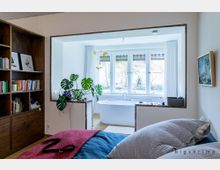 Schlafzimmer mit Blick in ein angrenzendes Badezimmer, das durch eine offene Durchreiche getrennt ist. Im Vordergrund ein Bett, daneben ein Regal mit Büchern, und im Badezimmer eine freistehende Badewanne vor einem großen Fenster mit Blick auf einen Garten.